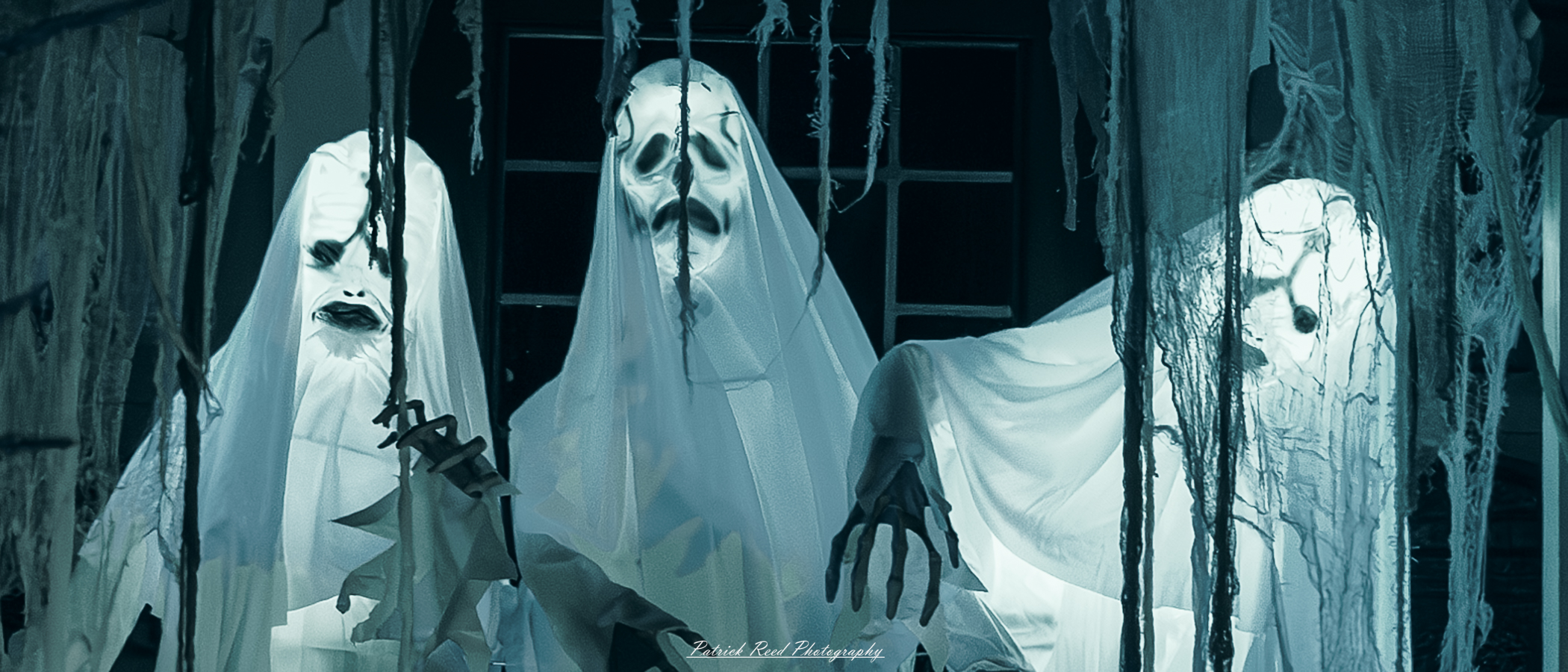 Taken on Tilson Street in Romeo Michigan on Halloween. Three ghostly figures appear in front of a dark, old-fashioned window, their eerie forms floating in misty white shrouds that almost blend with the shadows around them. Each ghost has hollow, empty eyes and faint, distorted facial features that seem to shift as you look at them. Their expressions are twisted and menacing, with wisps of ghostly fog curling around their forms. The room behind them is dimly lit, casting just enough light to reveal their translucent shapes, giving them a haunting, otherworldly glow. The atmosphere is heavy with dread, and the window is streaked with grime, adding to the unsettling mood.